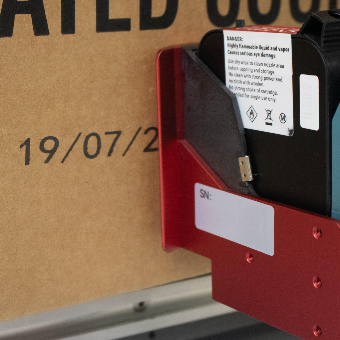 Detailed close-up of SNEEDJET print cartridge mounted in red print head assembly next to printed date code on cardboard