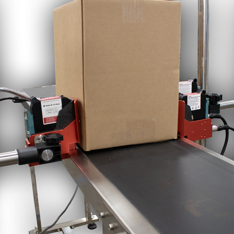 Close-up of industrial inkjet print heads applying a date code to a corrugated cardboard box on a conveyor system.